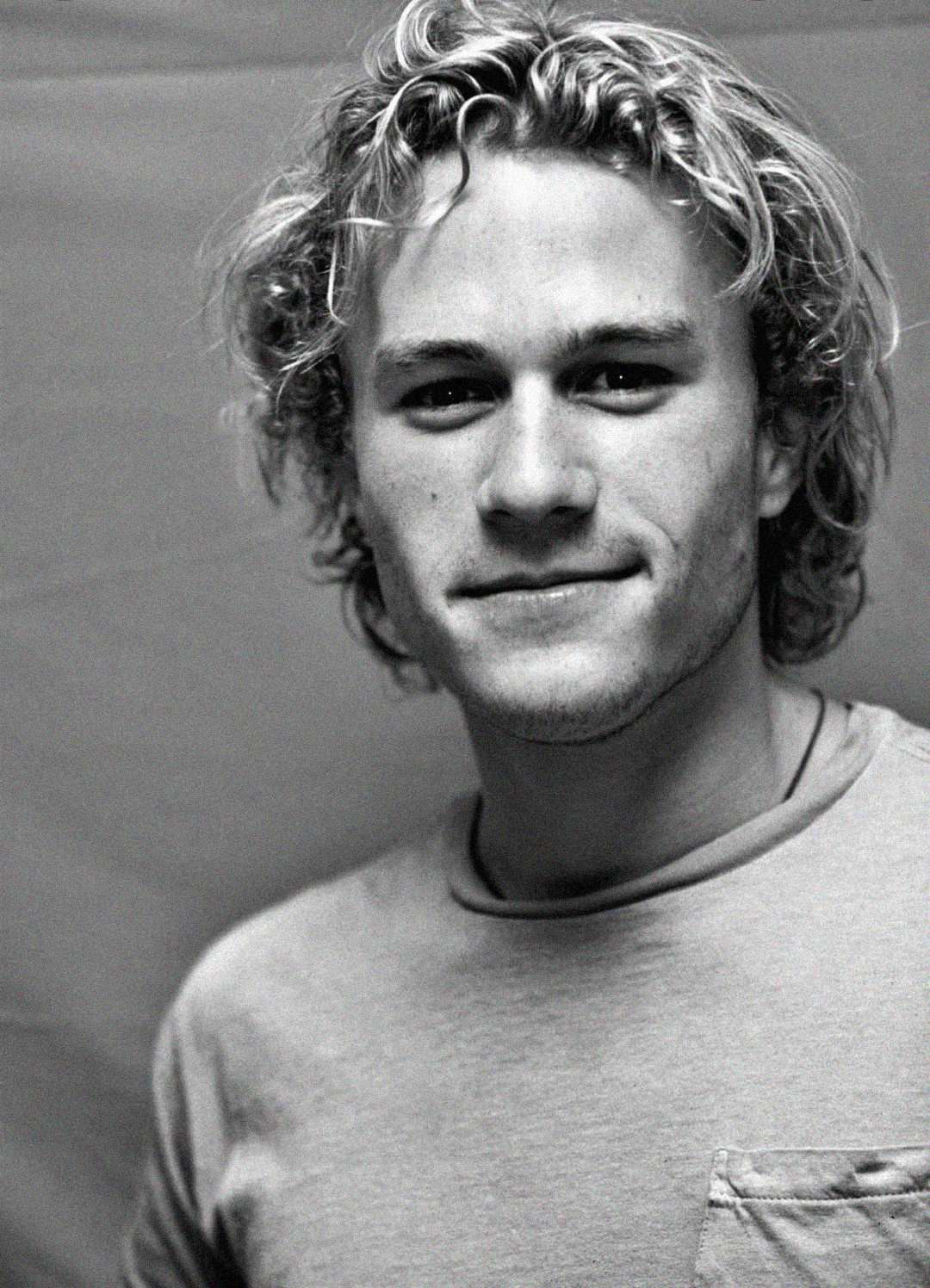 Un retrato en blanco y negro de un joven de cabello rubio ondulado, vestido con una camiseta de color claro y mirando a la cámara con una leve sonrisa. El fondo es sencillo y suavemente iluminado.