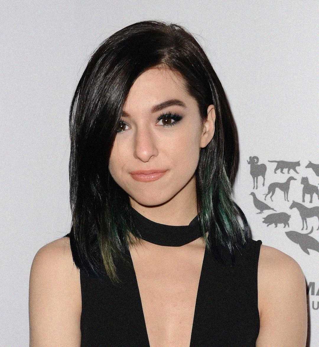 Una mujer con cabello liso y oscuro hasta los hombros y un maquillaje sutil viste un vestido negro sin mangas con escote tipo gargantilla, de pie frente a un fondo gris claro con siluetas de animales.