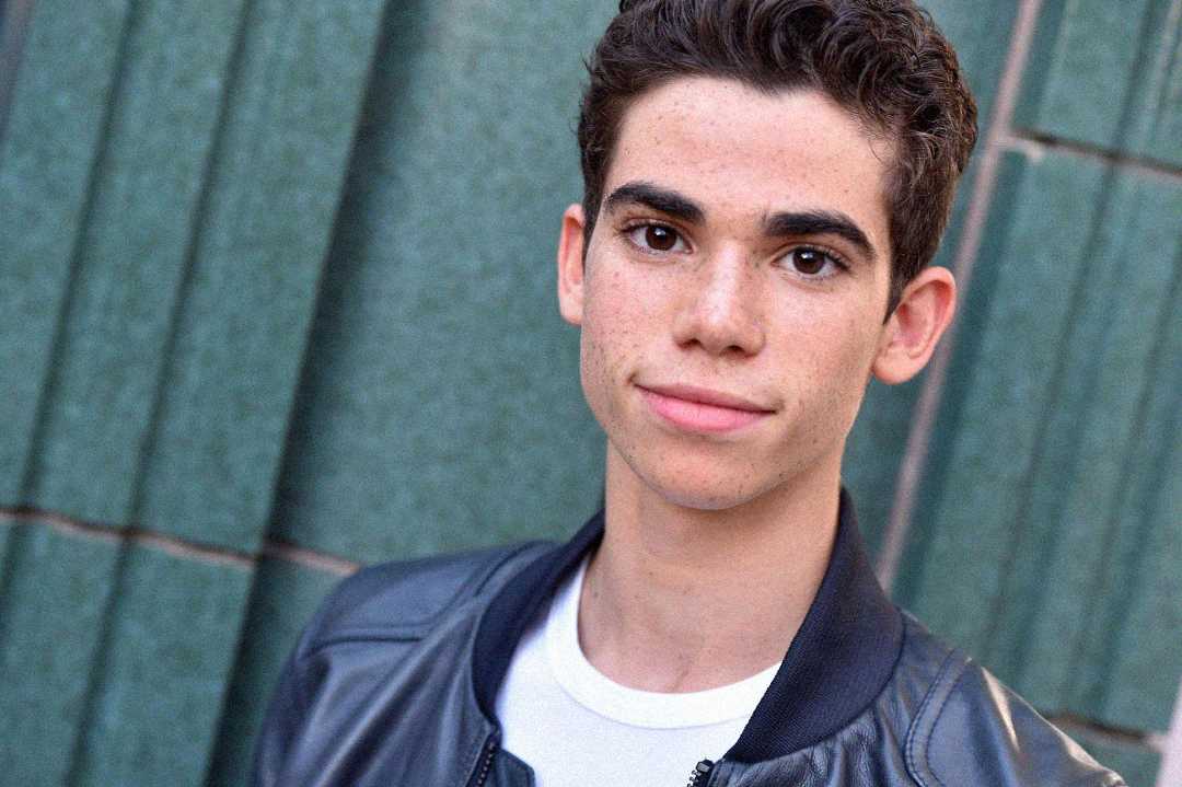 Un joven de pelo corto, rizado y oscuro, vestido con una chaqueta de cuero negra y una camisa blanca, se para frente a una pared de azulejos verdes, mirando a la cámara con una leve sonrisa.