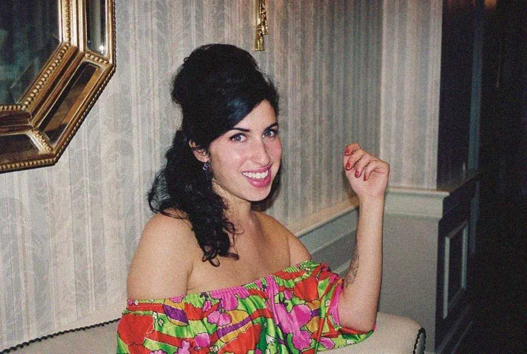 Una mujer sonriente con cabello oscuro peinado en forma de colmena se sienta en una silla blanca y lleva un top floral colorido con hombros descubiertos. El fondo muestra papel pintado estampado y un espejo con marco dorado.