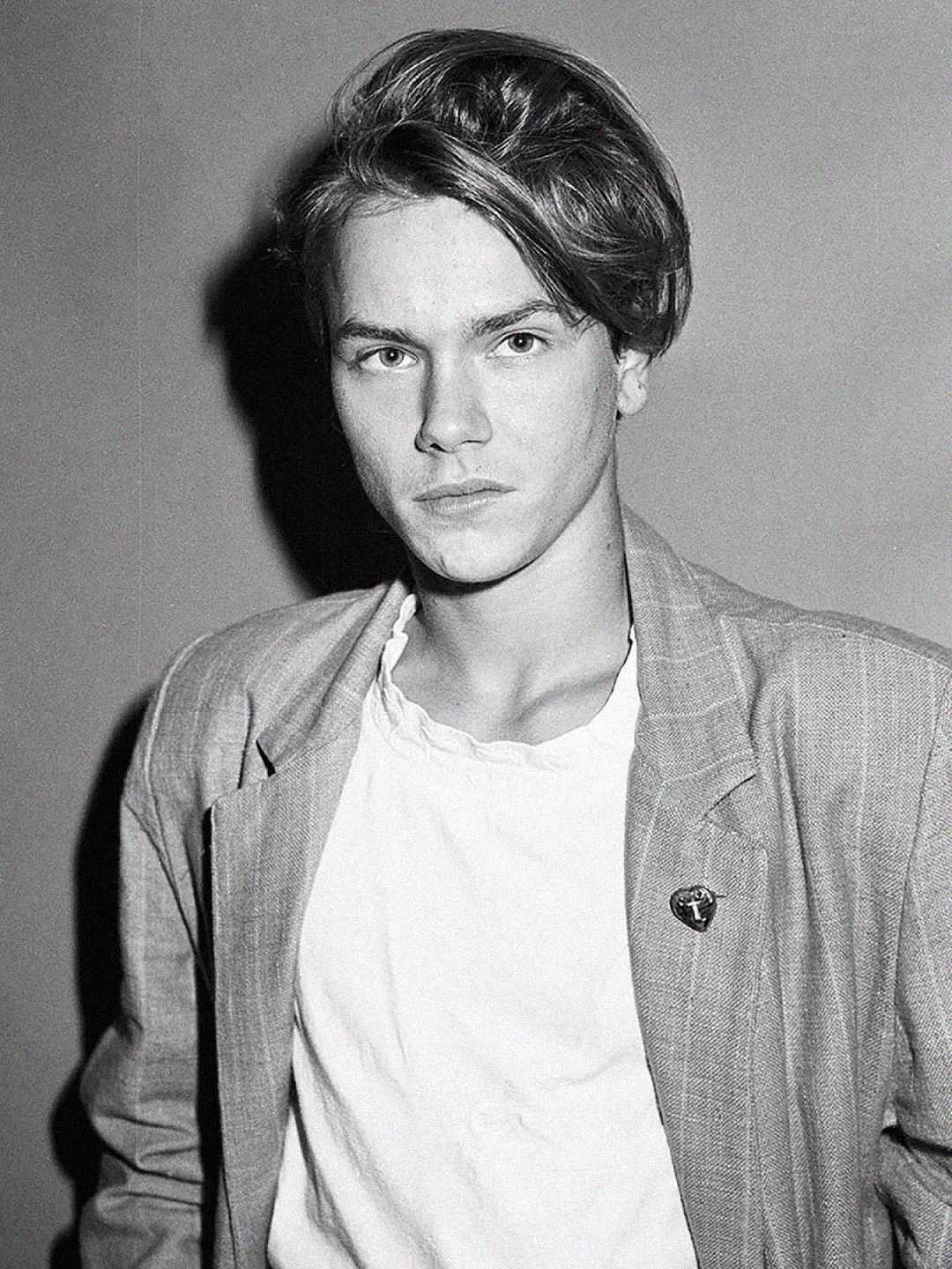 Fotografía en blanco y negro de un joven de pelo medio largo que viste un blazer de color claro sobre una camiseta blanca y mira seriamente a la cámara sobre un fondo liso.