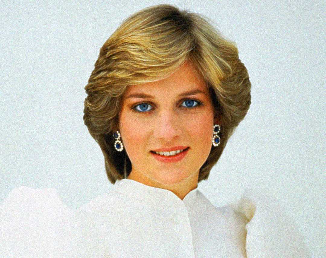 Una mujer de cabello corto, castaño claro y ojos azules sonríe suavemente. Lleva una blusa blanca con hombros abullonados y elegantes pendientes sobre un fondo liso y claro.