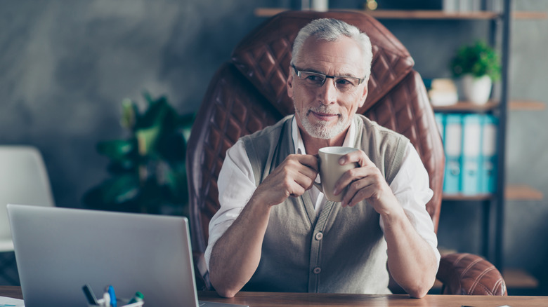 A senior-age professional smiles at his computer while holding a coffee mug