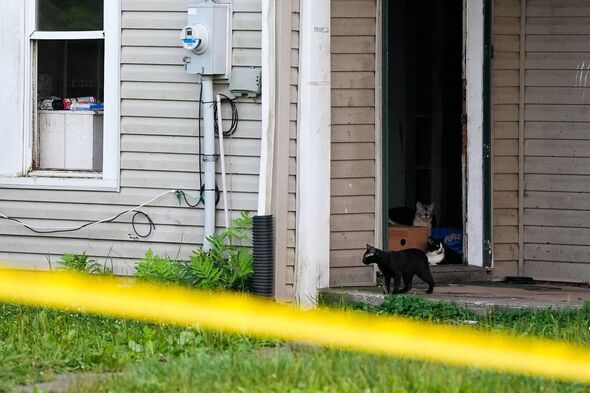 An individual is seated on the porch of a residential building, which is cordoned off with yellow tape, indicating an active inv