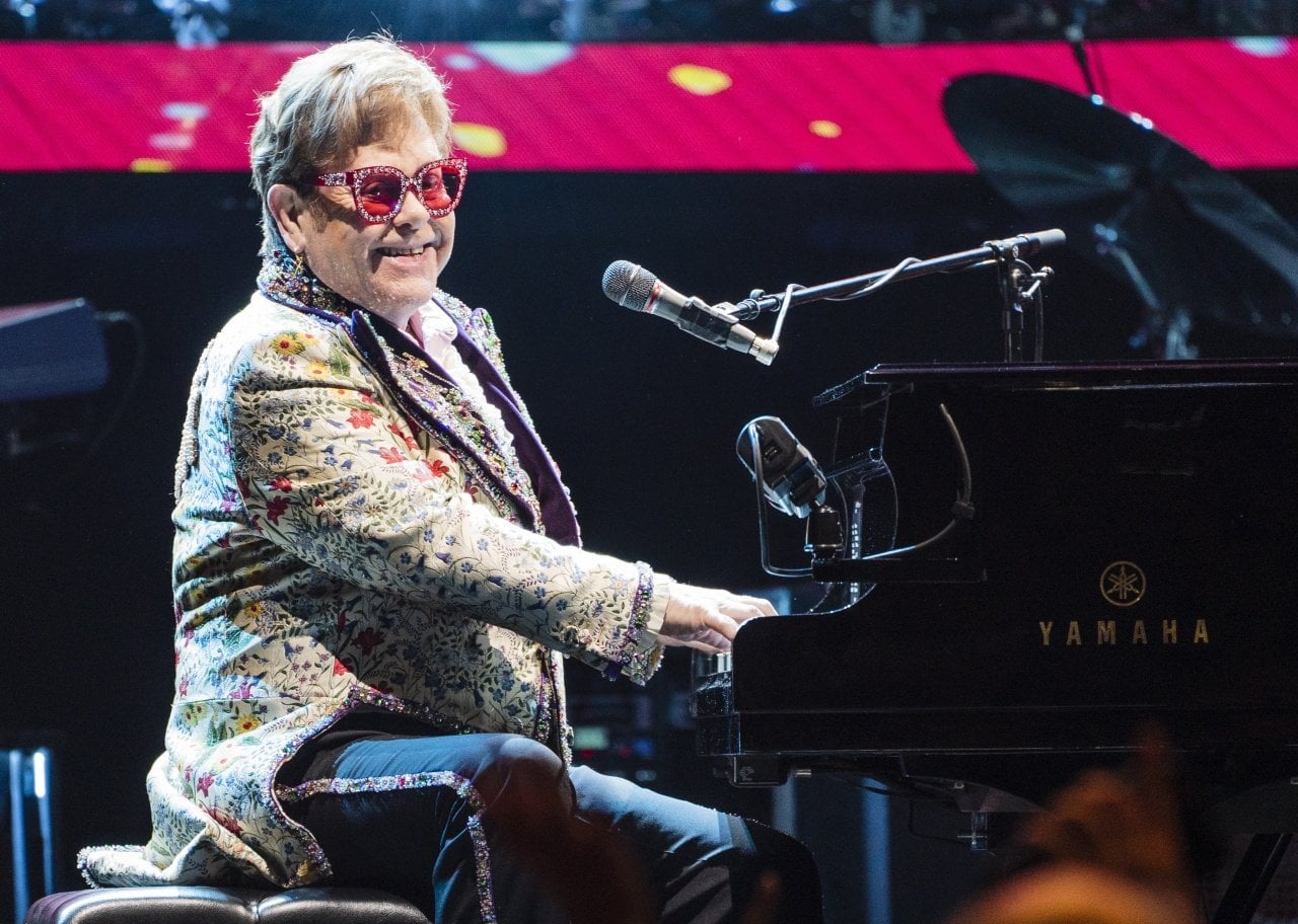 Elton John playing the piano in a floral jacket and sparkly sunglasses. - 