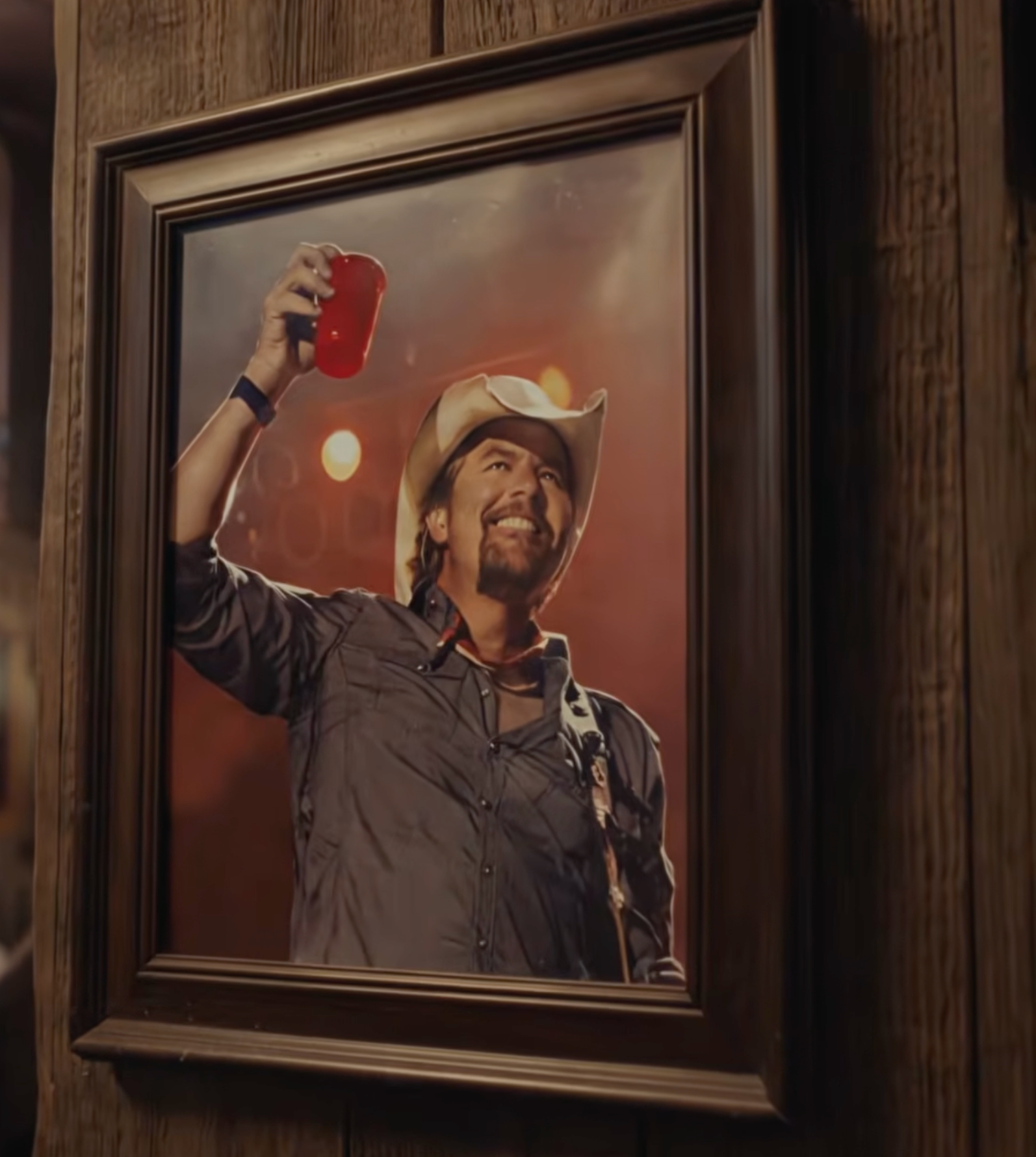 Un hombre con sombrero de vaquero anima con una taza roja en un retrato enmarcado sobre un fondo cálido y suavemente iluminado.