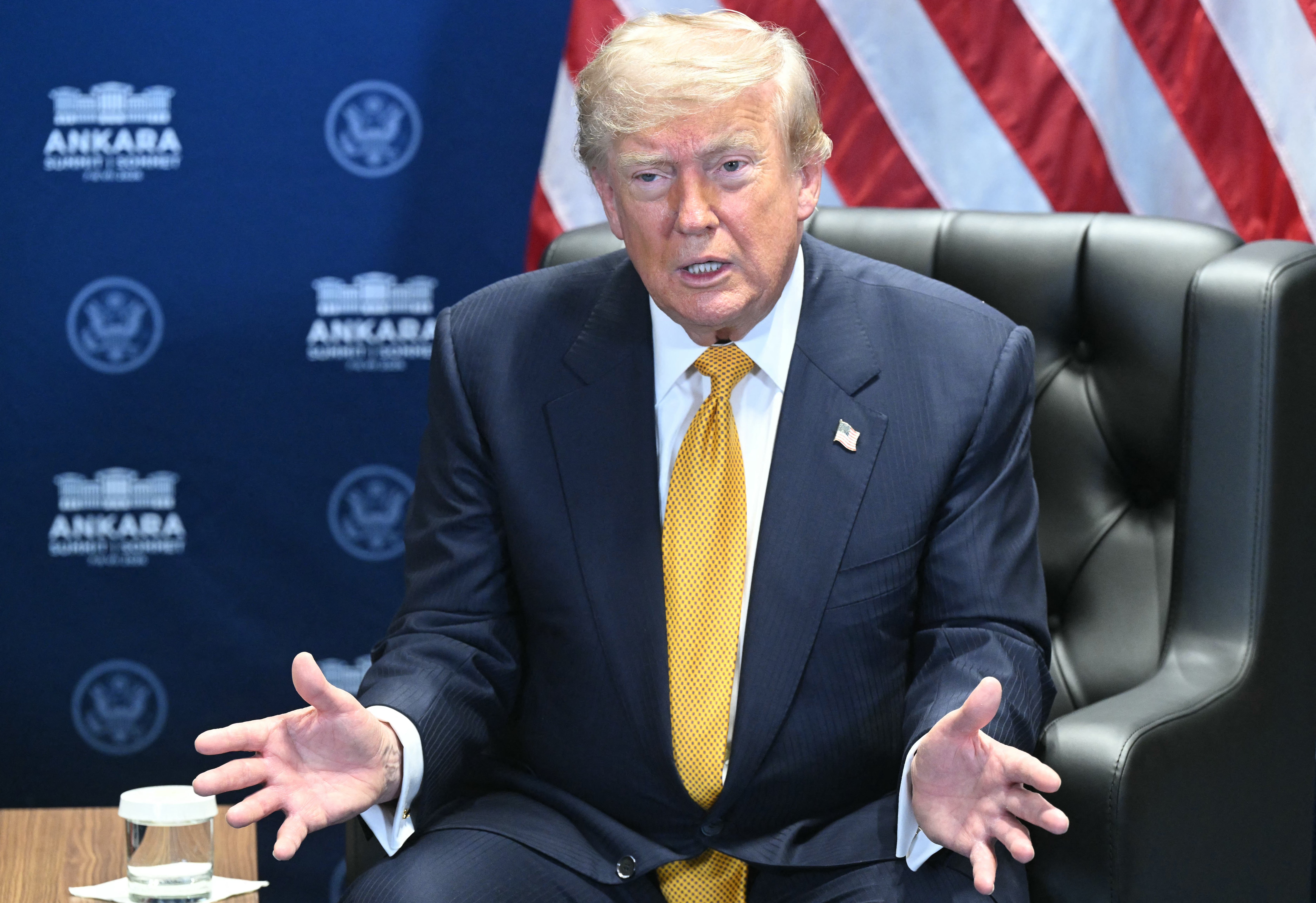 A man in a suit and tie is speaking while seated in front of a backdrop with flags and logos