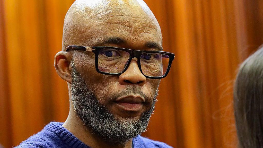 A head and shoulders picture of Cat Matlala in court - he is wearing a blue jumper, the neckline of which can just be seen at the bottom of the picture.