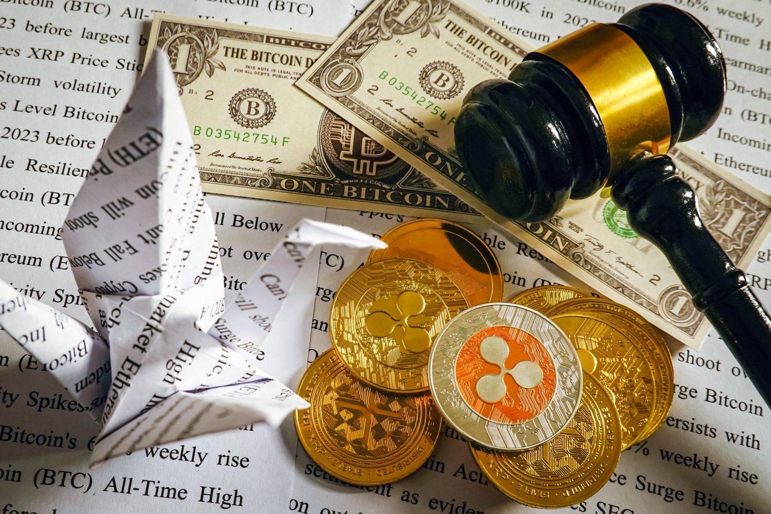 An overhead view features a wooden gavel resting on US dollar bills, which are printed with 'ONE BITCOIN'. Adjacent to these are a stack of gold-colored cryptocurrency coins, topped by a distinctive silver and orange Ripple (XRP) coin. In the foreground, an origami crane, folded from newspaper print containing financial terms like 'Bitcoin' and 'ETH', is visible. The entire arrangement is set against a background of blurred financial article text.