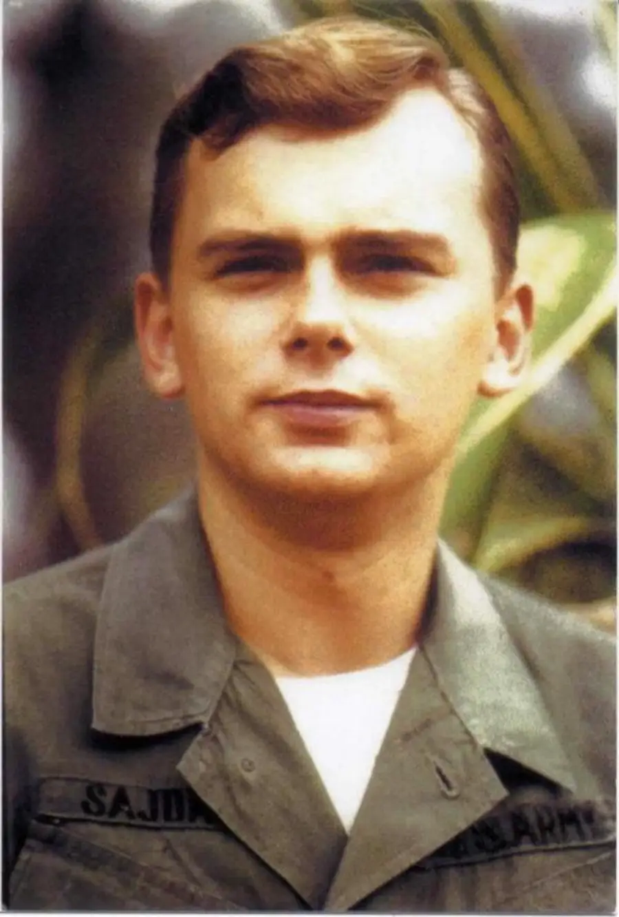 A young man in a green military uniform stands outdoors. His short, light brown hair is neatly styled, and he looks ahead with a neutral expression. The background is softly blurred with greenery.