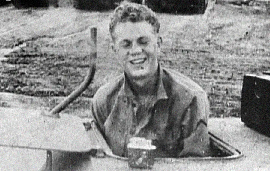 A young man in military uniform smiles while emerging from a tank hatch, with a cup placed on the tank near him. The photo is black and white, and the background shows a paved ground area.