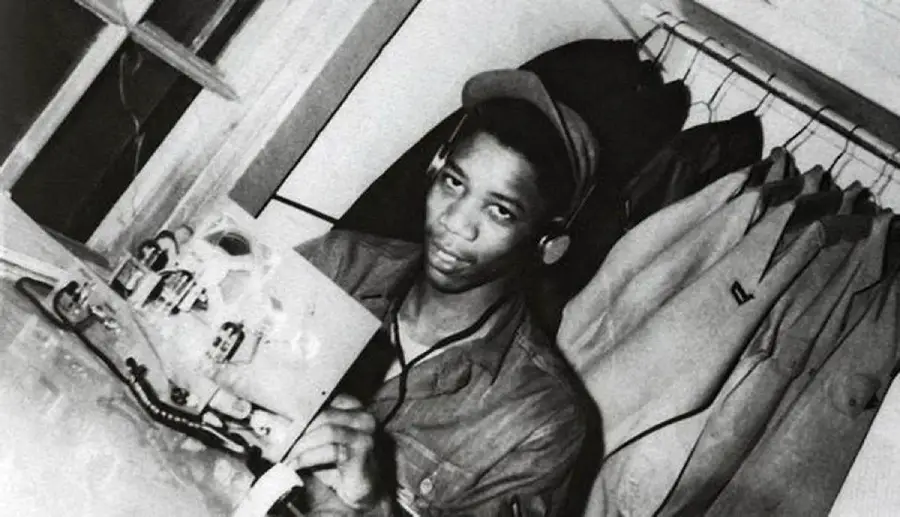 A Black man in military uniform sits indoors, holding electronic equipment with headphones on. Coats hang behind him. He looks toward the camera.