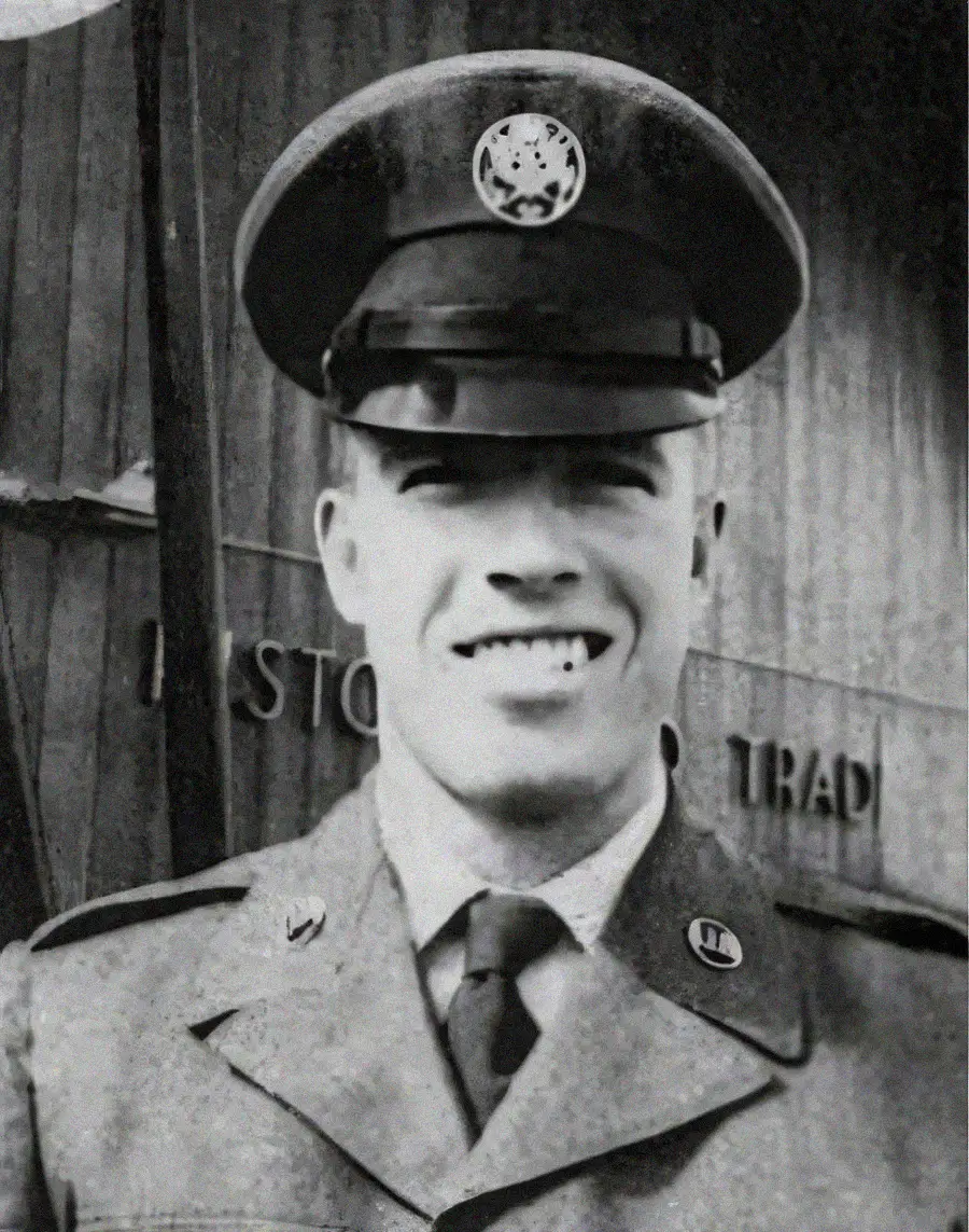 A smiling man in a military uniform and cap stands outside, with a building and partially visible sign in the background. The photo is black and white.