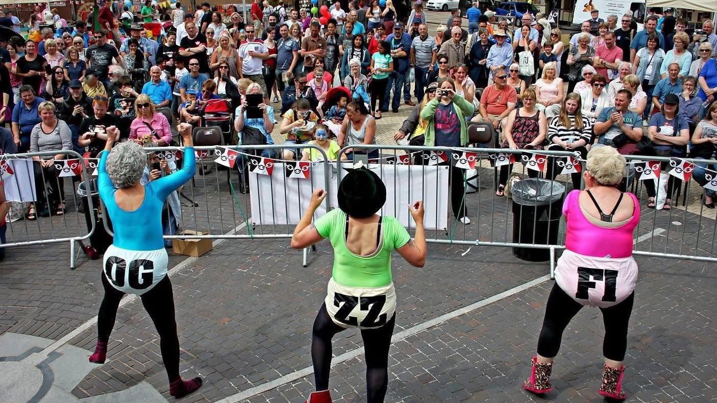 Three women are standing in front of a crowd on a high street, wearing neon coloured unitards and blonde curly wigs. They have pointed their hands in the air and are dancing. 