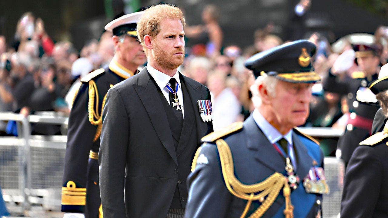 El rey Carlos caminando con un uniforme azul y luciendo sombrío frente al príncipe Harry con un traje oscuro.