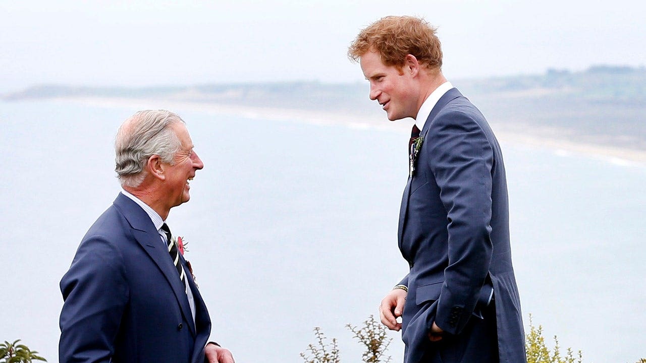 El rey Carlos y el joven príncipe Harry, vestidos con trajes azul oscuro a juego, conversan y sonríen.