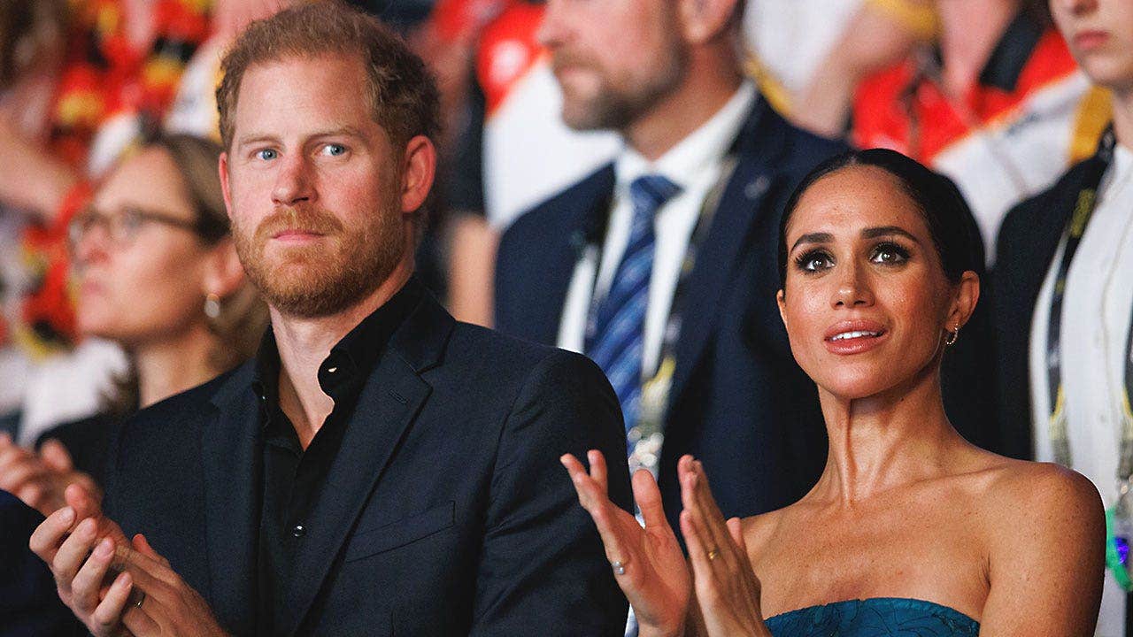 El príncipe Harry y Meghan Markle están uno cerca del otro en una audiencia aplaudiendo y luciendo serios.