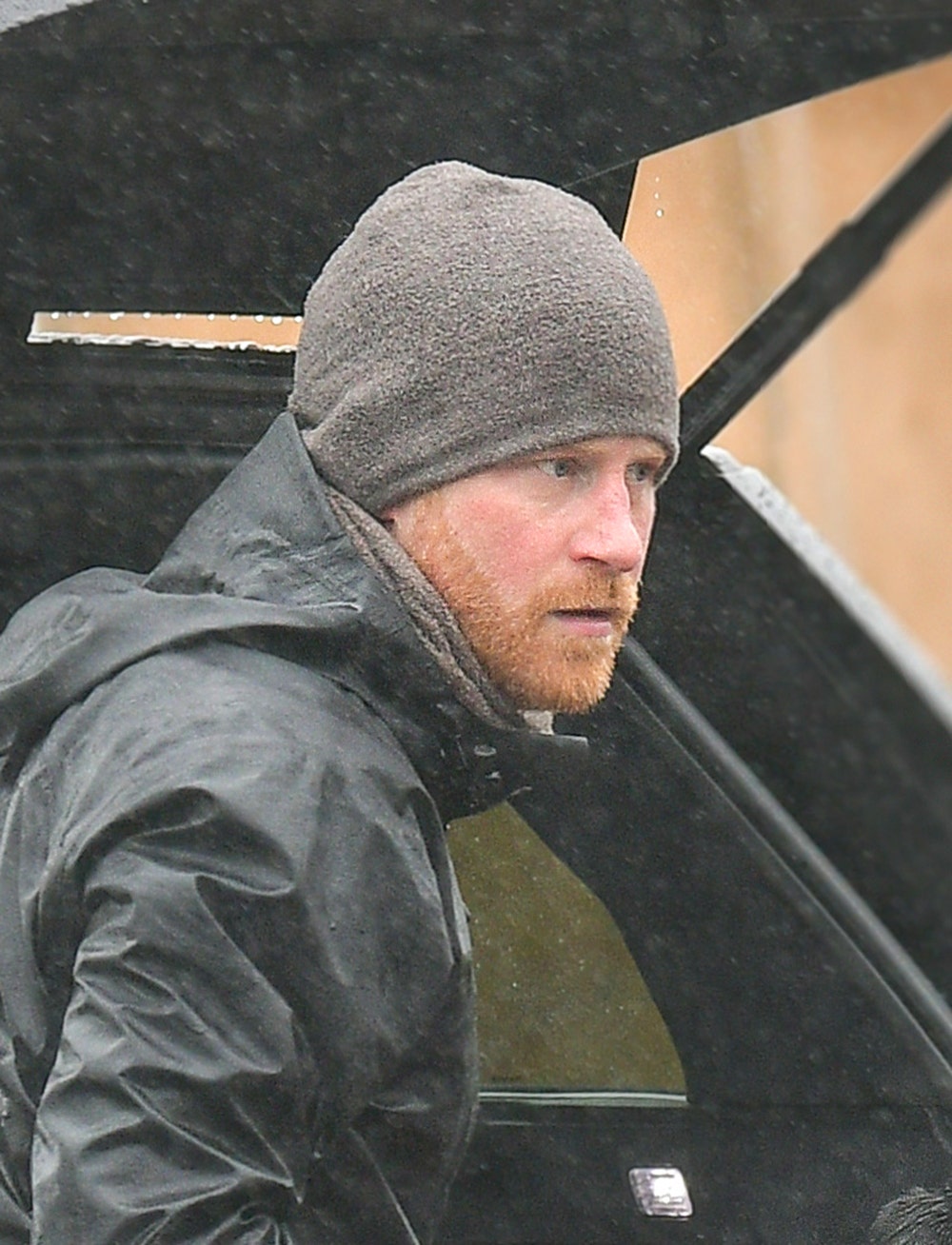 El Príncipe Harry con una chaqueta gris y un gorro a juego parado frente a un auto en Montecito luciendo serio.