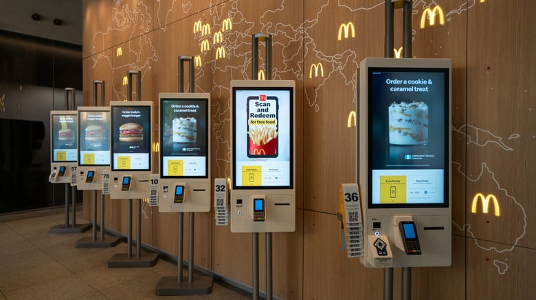 A row of self-service digital kiosks at a McDonald's restaurant, displaying menu items and promotions, with card readers and order numbers in front of a wall decorated with a world map and McDonald's logos.