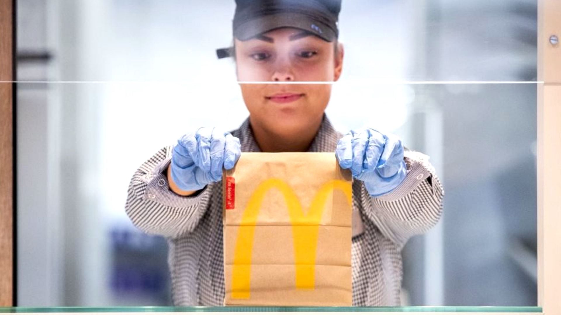 A McDonald's employee wearing a uniform, cap, and blue gloves hands a brown paper bag with a yellow McDonald's logo to a customer across a counter.