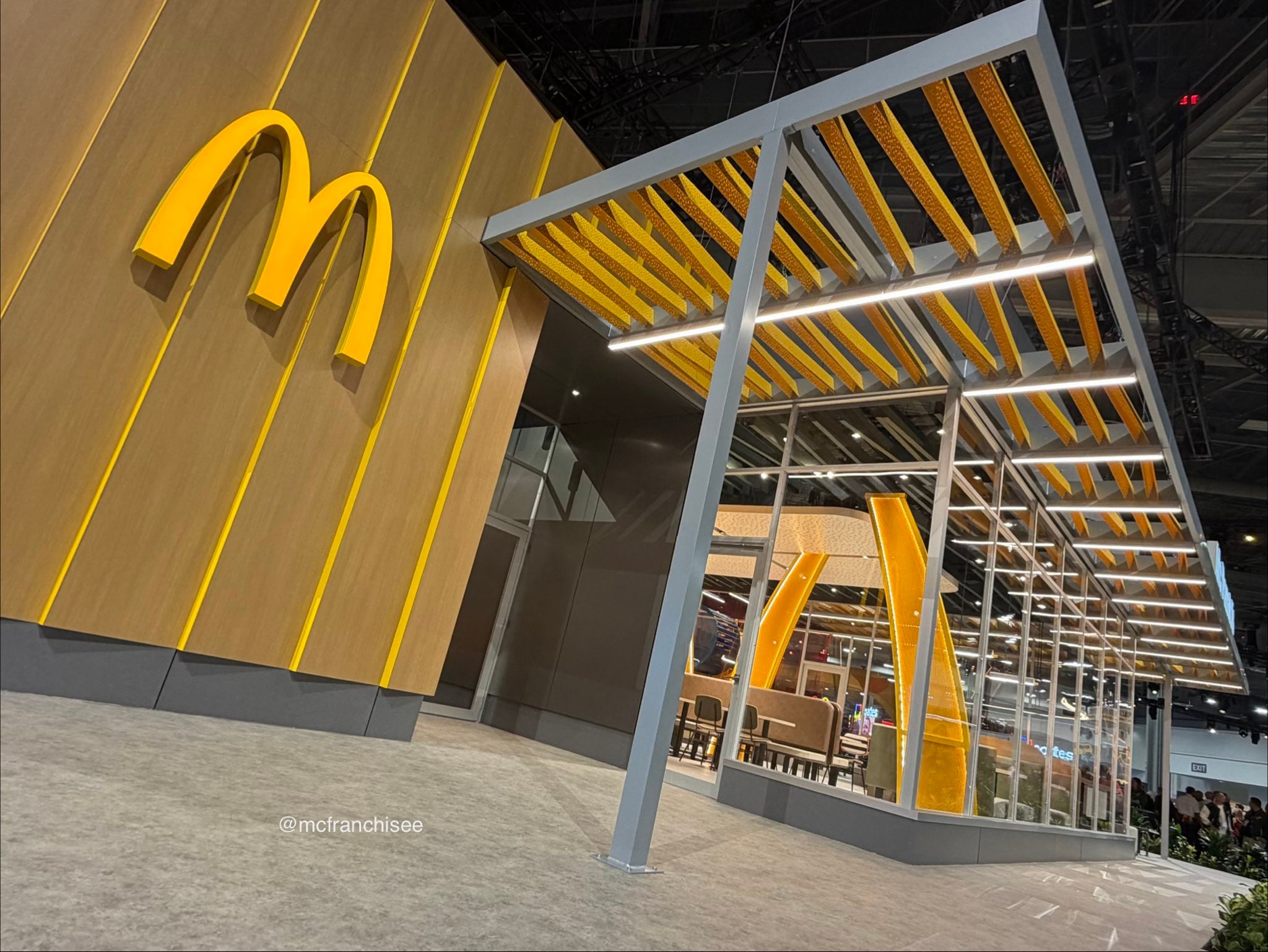 A modern McDonald’s restaurant exterior with yellow arches and a unique yellow-striped metal canopy over an outdoor seating area, viewed from a low angle indoors.