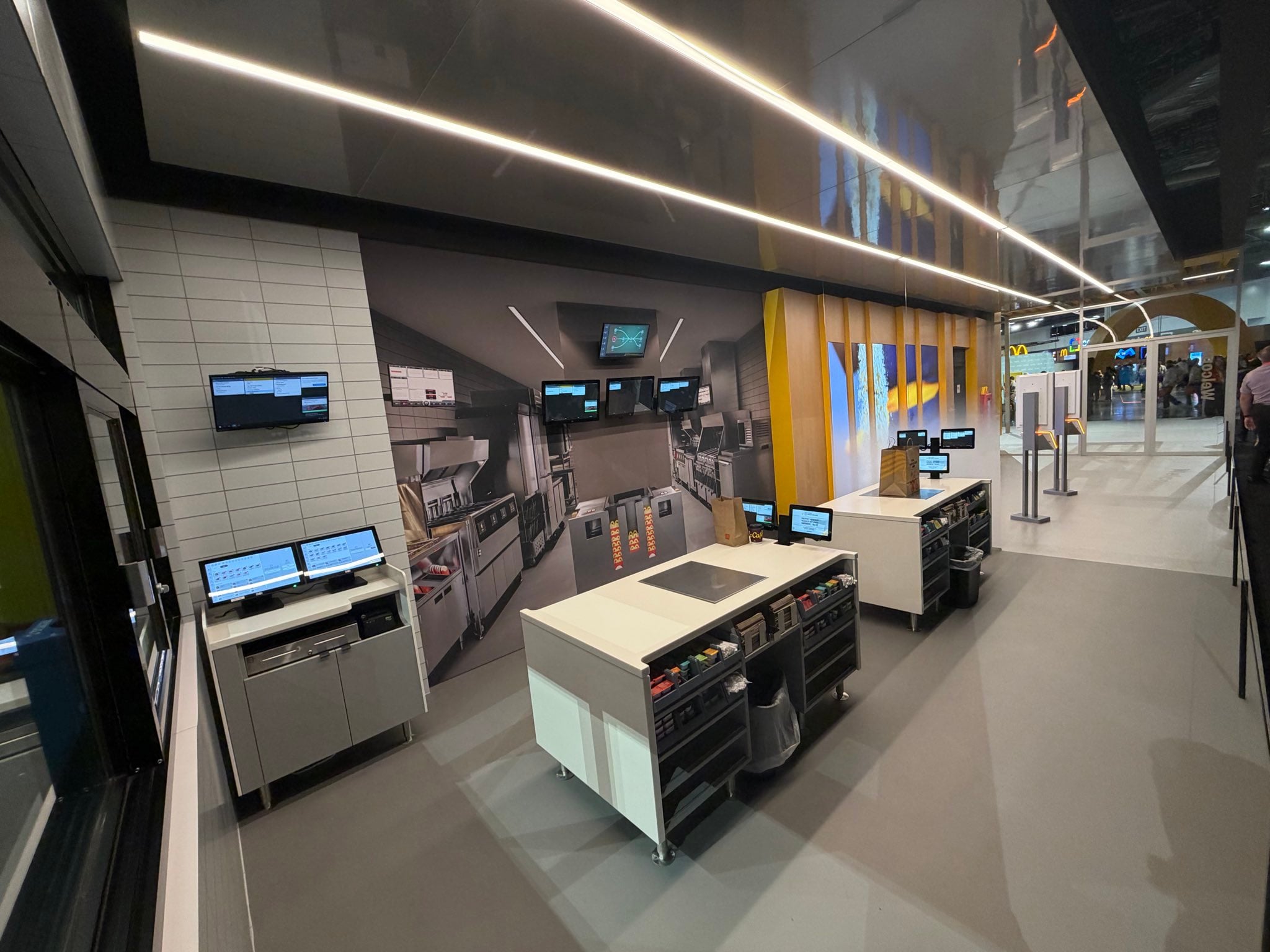 A modern fast-food kitchen with self-service kiosks, order screens, and stainless steel equipment. Yellow accents and a mural are on the wall. People wait in line in the background.