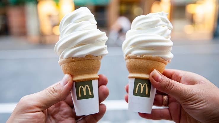McDonald's foods. Two hands holding McDonald's vanilla soft serve ice cream cones with the logo visible, against a blurred outdoor background.