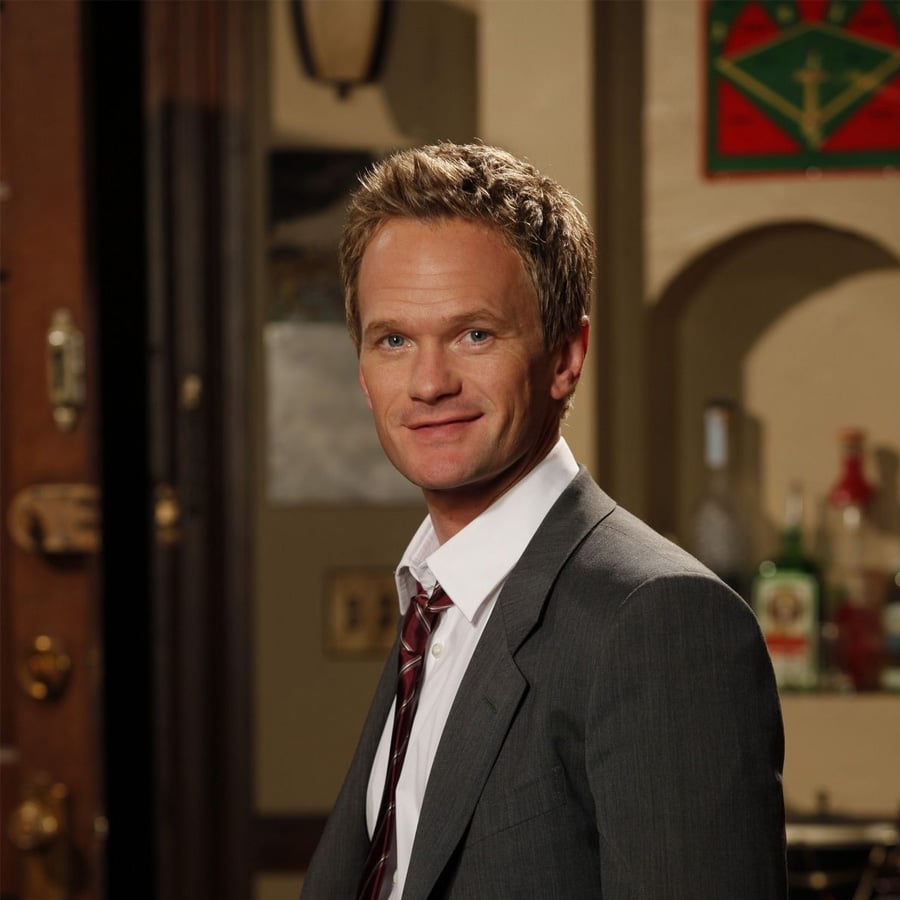 A man in a gray suit and loose tie stands indoors, smiling slightly. Shelves with bottles and a colorful wall hanging are visible in the background, giving a casual setting.