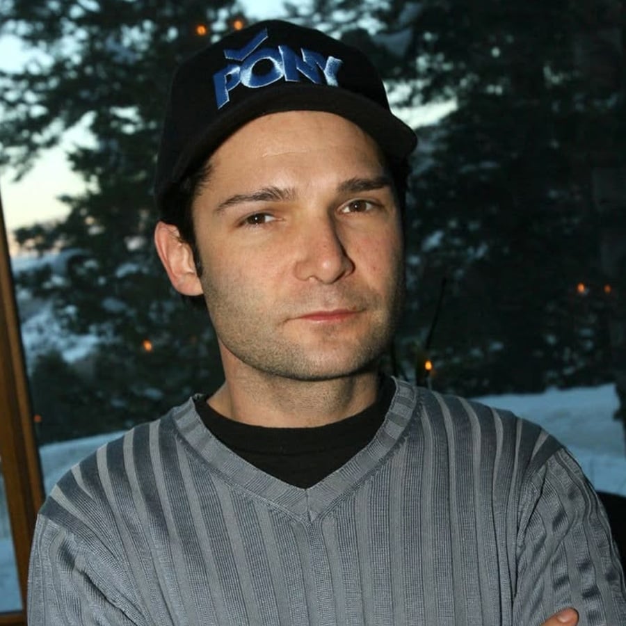 A person wearing a black "PONY" cap and a gray striped sweater stands indoors with arms crossed, with a snowy outdoor scene visible through the window behind them.