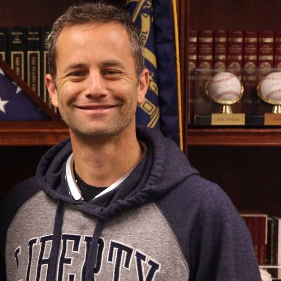 A smiling man in a "Liberty" hoodie stands in front of a bookshelf with books, baseballs in display cases, and a partially visible flag.