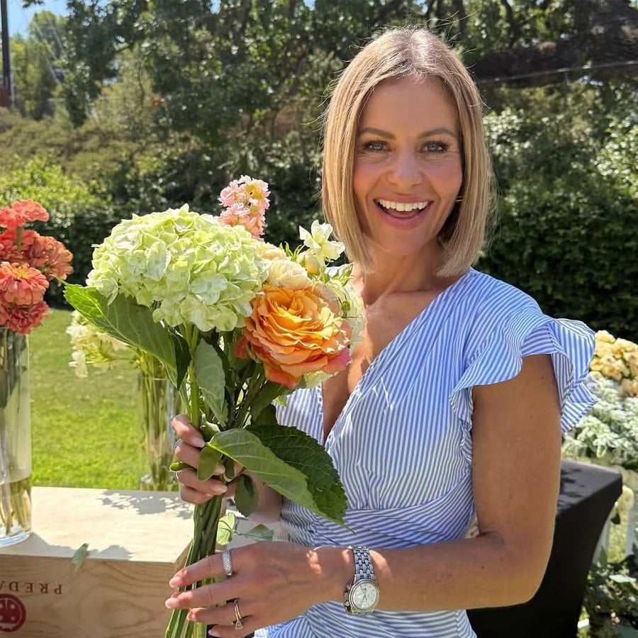 A smiling woman in a blue and white striped dress holds a bouquet of hydrangeas and roses outdoors, with more flowers and greenery in the background on a sunny day.