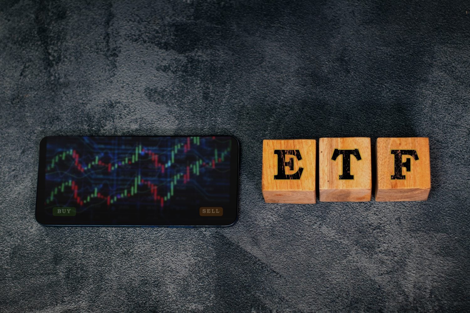 A smartphone displaying a financial candlestick chart with green and red lines, and 'BUY' and 'SELL' buttons at the bottom. To the right of the phone are three light brown wooden blocks, each with a single black capital letter, spelling 'E', 'T', 'F' from left to right. The background is a textured dark grey surface.