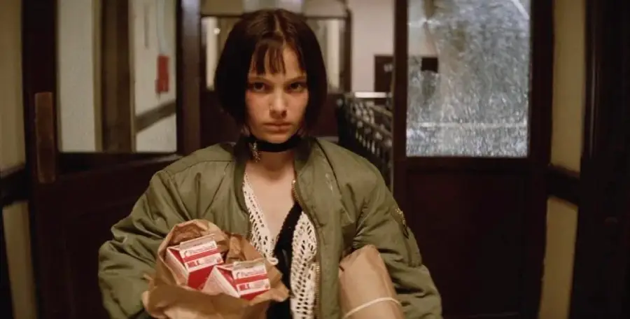A young girl with short dark hair in a green jacket carries grocery bags, one filled with milk and cartons, walking through broken glass doors in a dimly lit hallway.