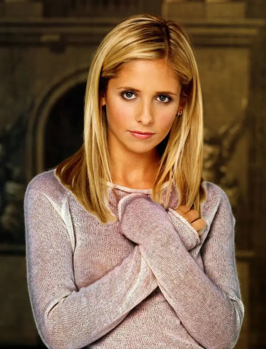 A woman with straight blonde hair and a serious expression wears a light purple sweater, standing indoors with a blurred, ornate background behind her.