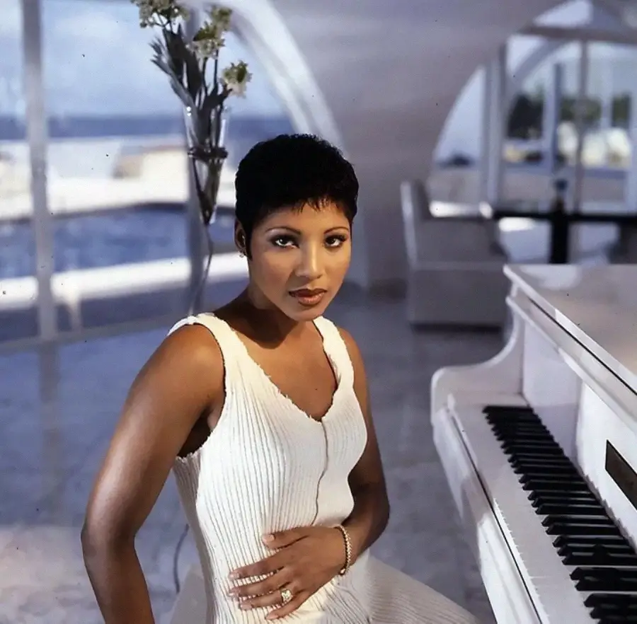 A woman in a white dress sits at a white grand piano in a modern, sunlit room with large windows and flowers in the background.