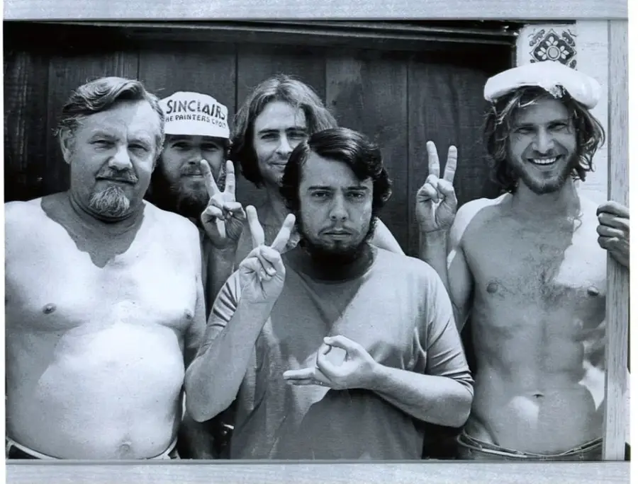 Five men pose together outside in a black-and-white photo. Three of them flash peace signs, one smiles while wearing a hat, and another man in front makes a different hand gesture. Most are shirtless and appear relaxed.