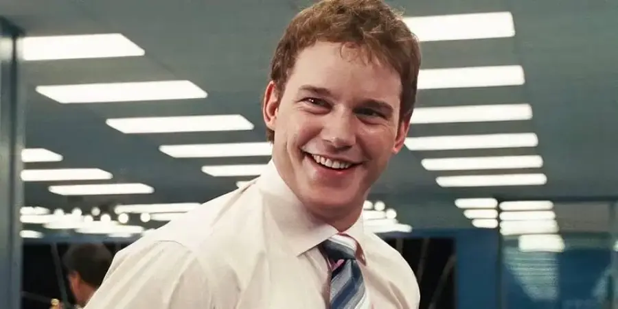 A man in a white shirt and striped tie smiles while standing indoors under bright ceiling lights.