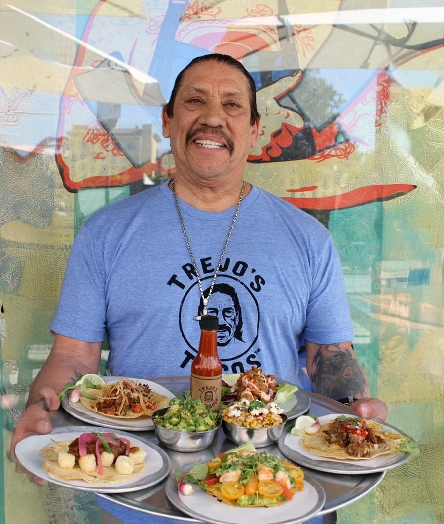 Um homem sorridente em um "Tacos do Trejo" camisa segura uma bandeja com tacos, guacamole e molho picante, em frente a um mural colorido.