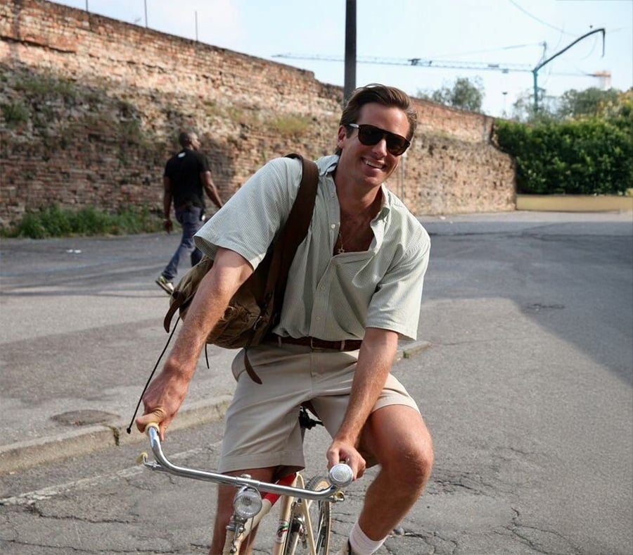 Um homem sorridente usando óculos escuros, camisa leve e shorts anda de bicicleta em uma rua ensolarada com uma parede de tijolos ao fundo.
