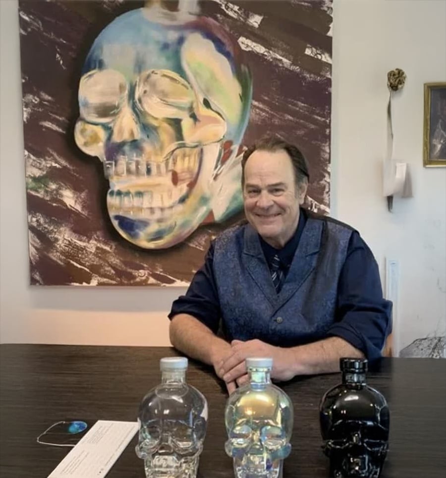 Um homem de colete azul está sentado a uma mesa com três garrafas em forma de caveira à sua frente. Atrás dele está pendurada uma pintura colorida de uma caveira na parede.