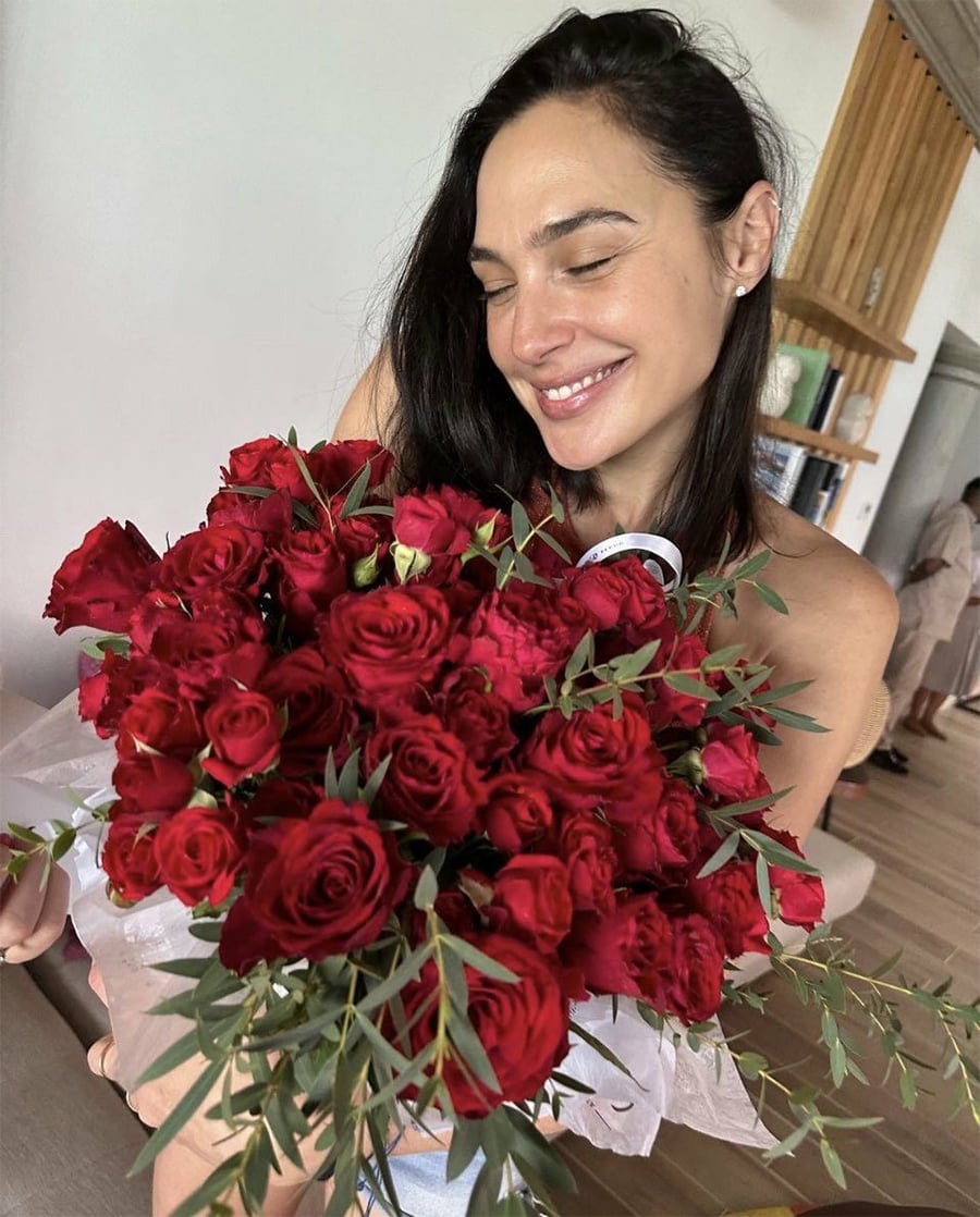 Uma mulher de cabelos escuros sorri enquanto segura um grande buquê de rosas vermelhas com folhagens. Ela está dentro de casa e prateleiras e outra pessoa são visíveis ao fundo.
