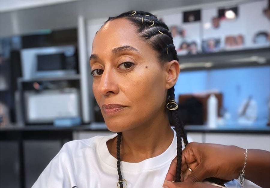 Uma mulher com tranças trancinhas, adornada com punhos dourados, está sentada em um salão enquanto alguém penteia seu cabelo. Ela veste uma camisa branca e olha com confiança para a câmera. O fundo mostra equipamentos e fotos do salão.