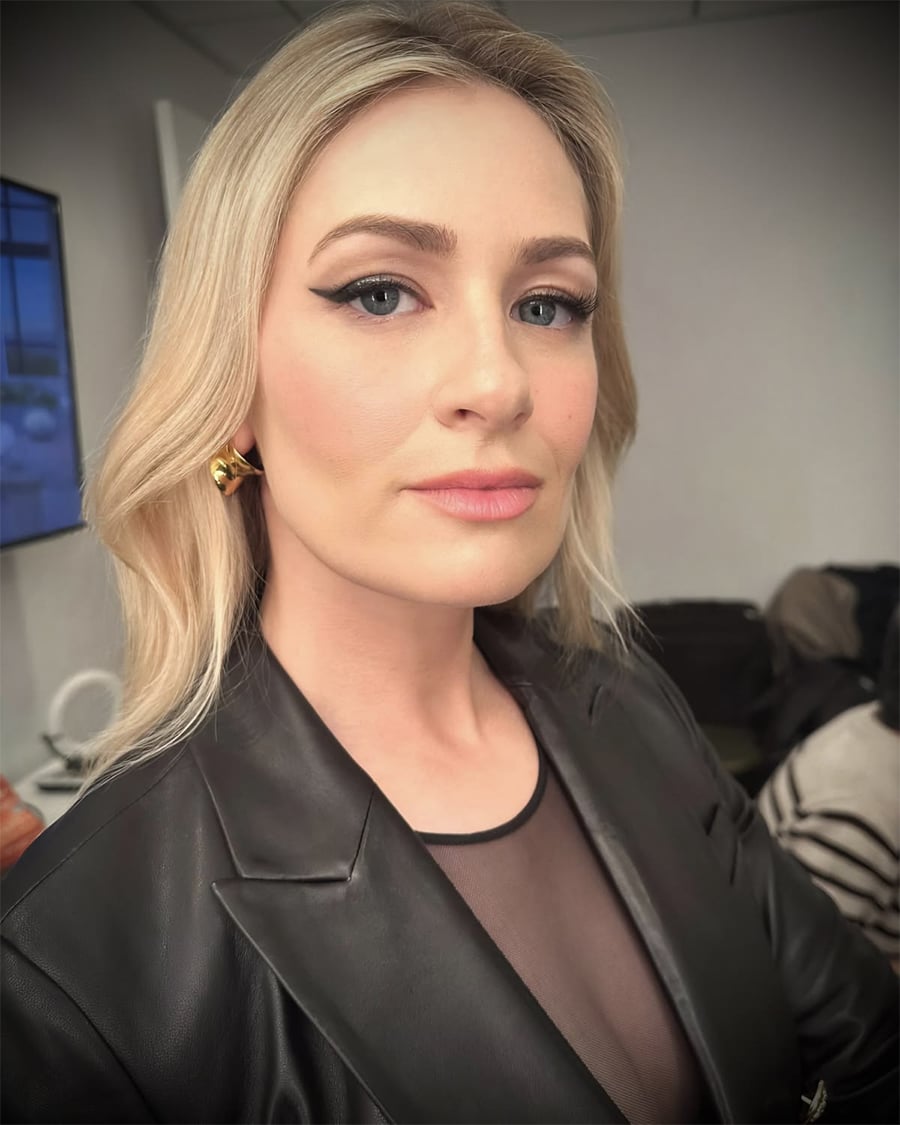 Uma mulher com longos cabelos loiros, vestindo um blazer preto e brincos dourados, olha com confiança para a câmera em um ambiente interno bem iluminado.
