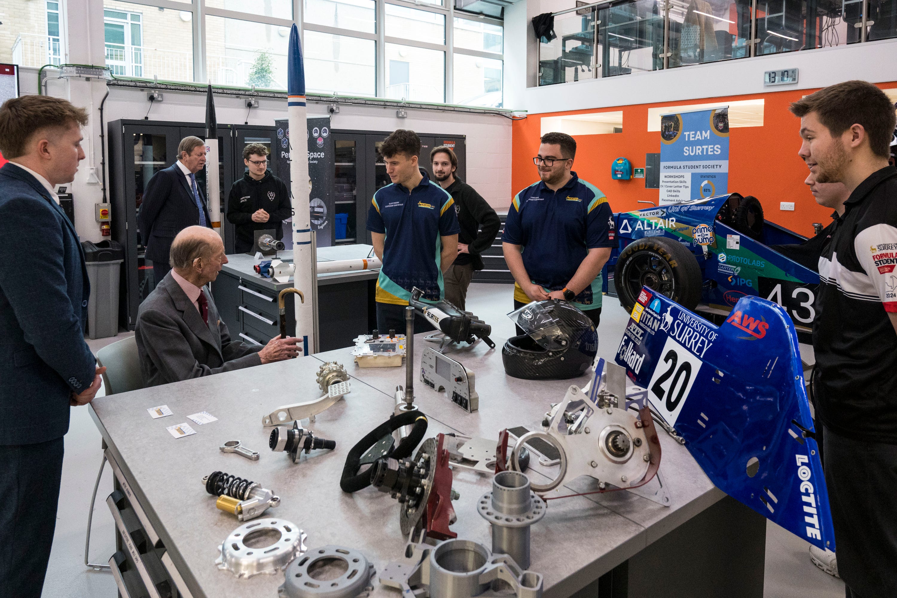 Group of students and a senior gentleman discussing engineering projects at a workspace with various models and components.