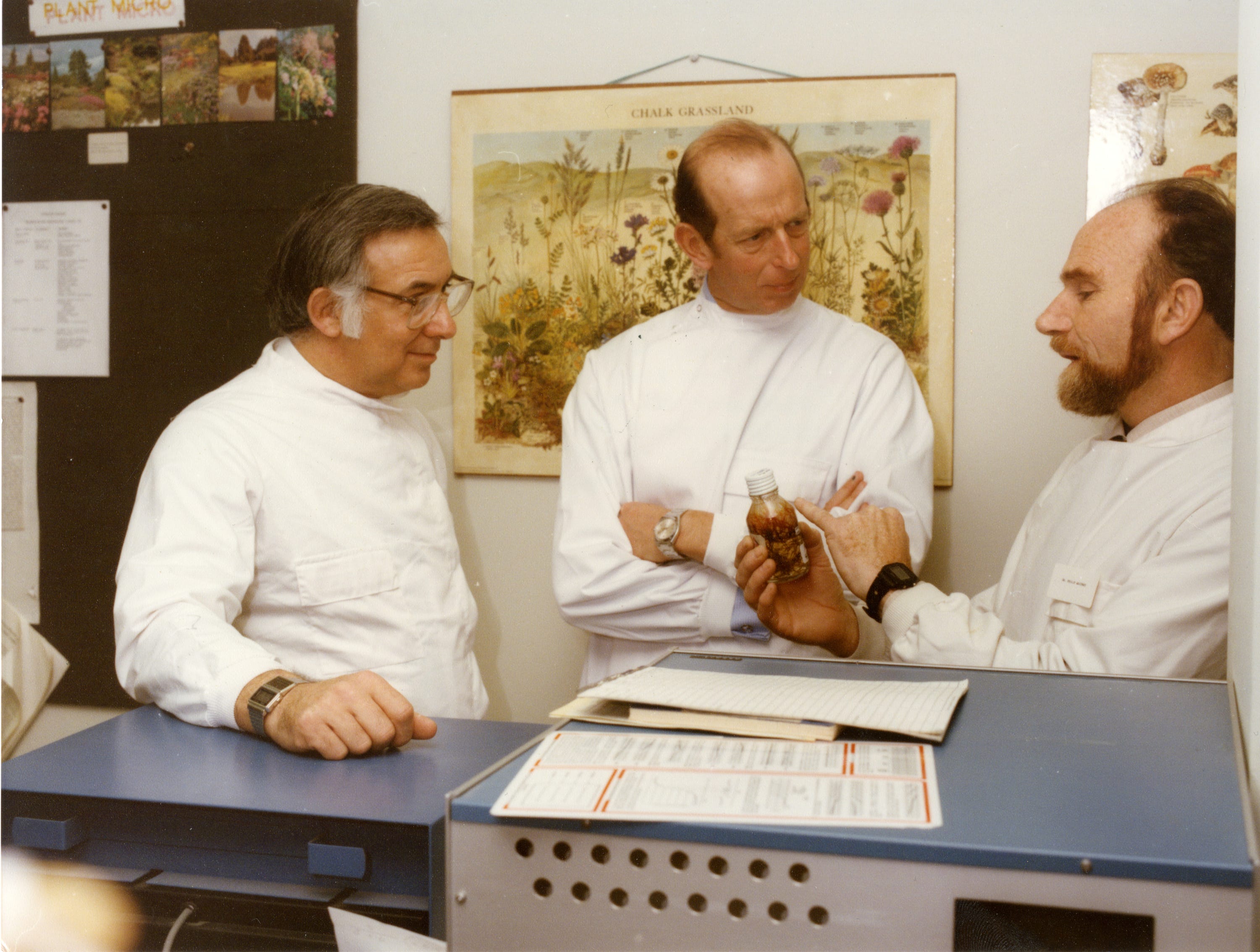 Three men in lab coats discuss a small bottle while standing in a laboratory environment with charts on the wall.