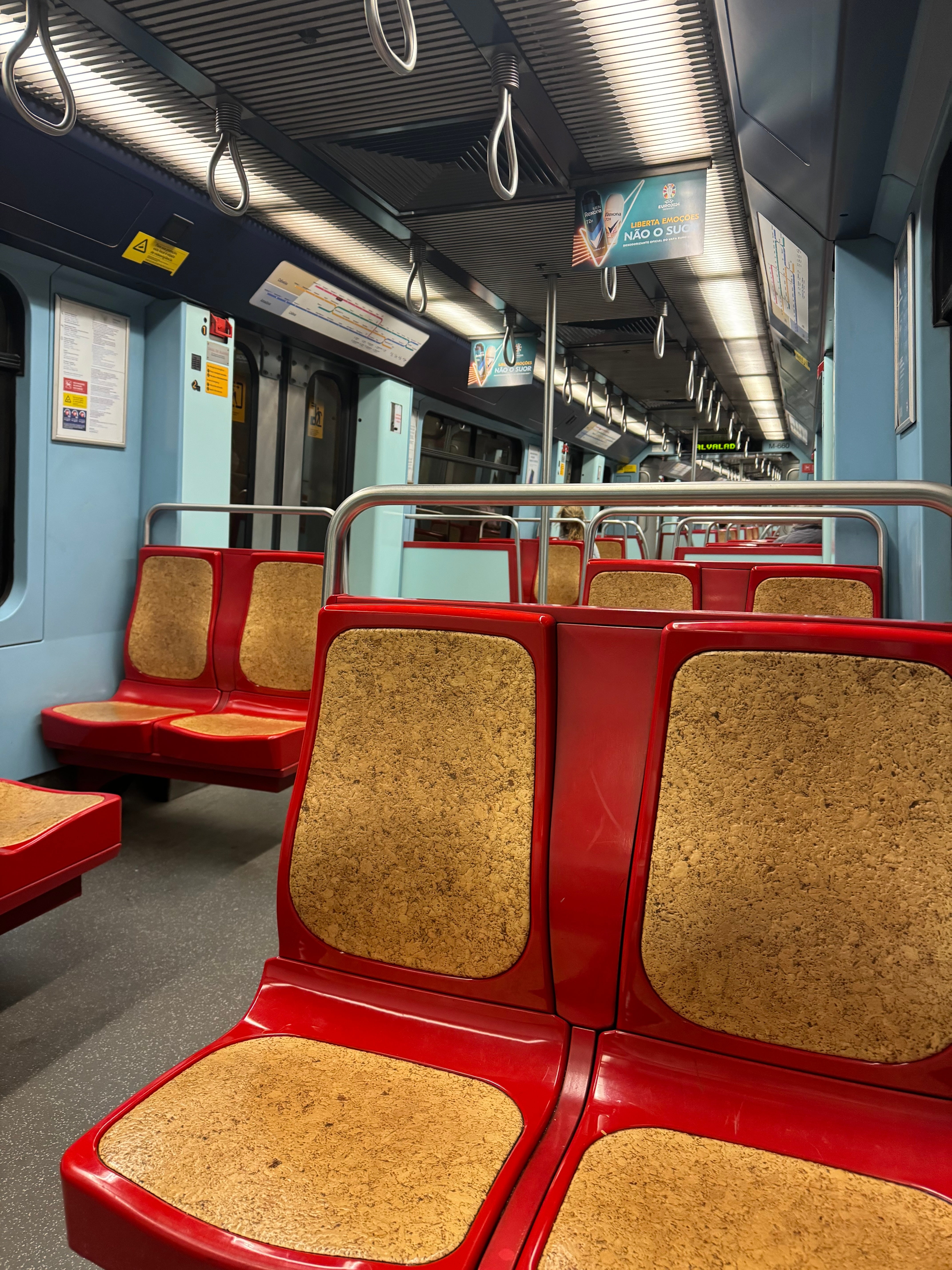 Vagão de metrô vazio com assentos almofadados vermelhos e corrimãos metálicos. Sinais suspensos e anúncios são visíveis