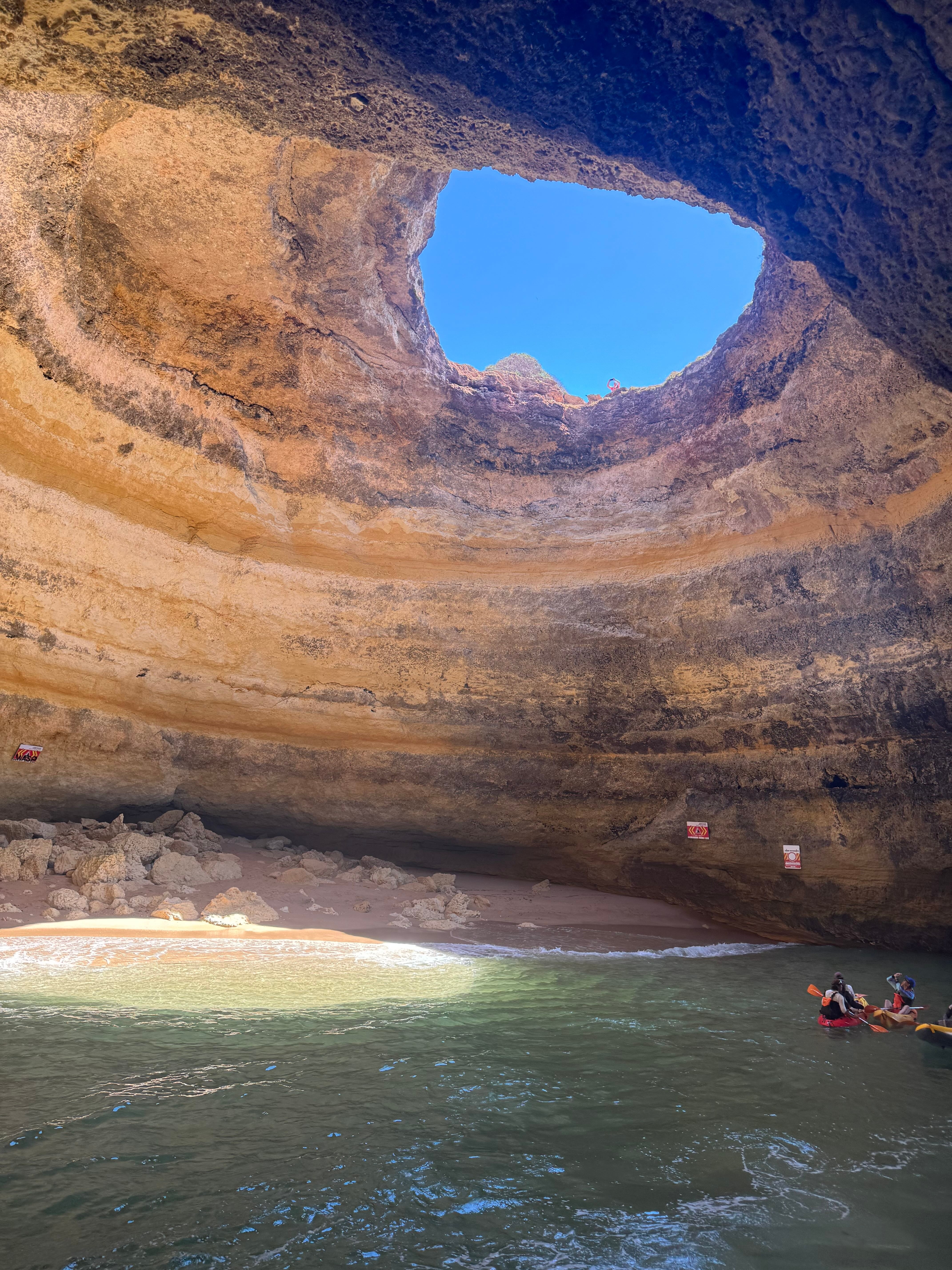 Os praticantes de caiaque exploram uma caverna marítima com uma grande abertura circular no topo, revelando o céu, e uma praia arenosa no interior