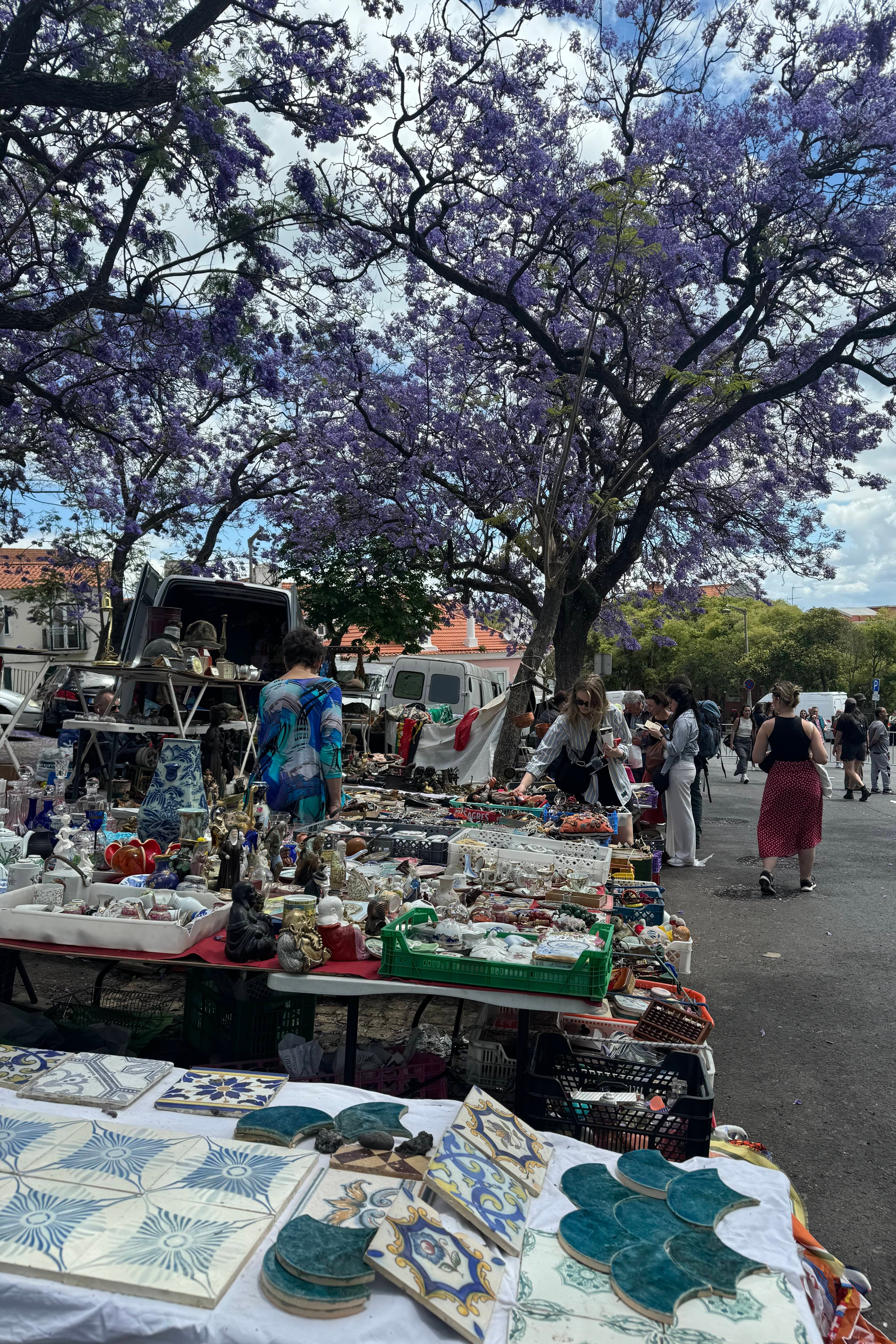 Mercado ao ar livre com visitantes examinando diversos produtos em mesas, à sombra de árvores floridas