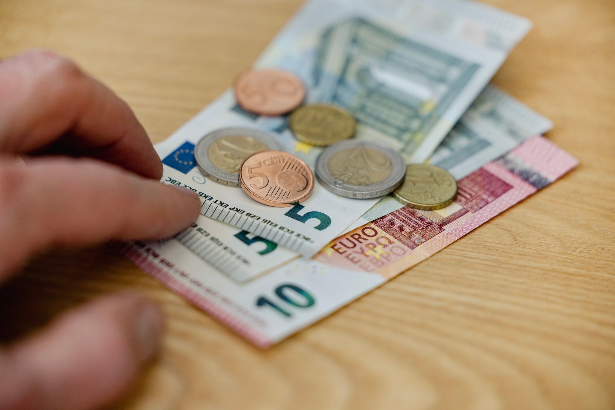 Várias notas e moedas de euro numa mesa de madeira com uma mão a alcançá-las, indicando um contexto financeiro relacionado com viagens