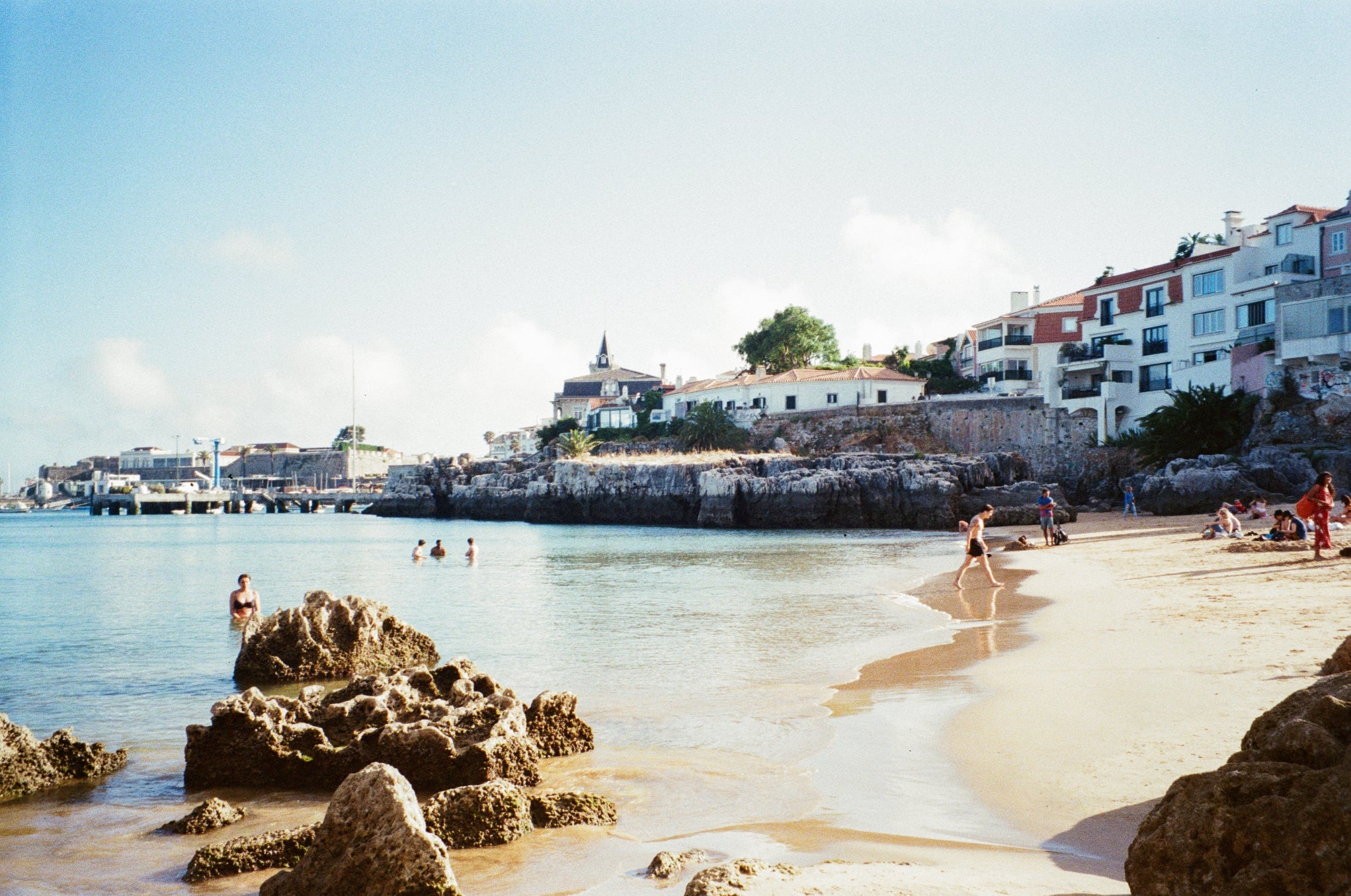 Praia com costa rochosa, águas claras, pessoas nadando e tomando sol e casas ao longo da costa sob um céu claro